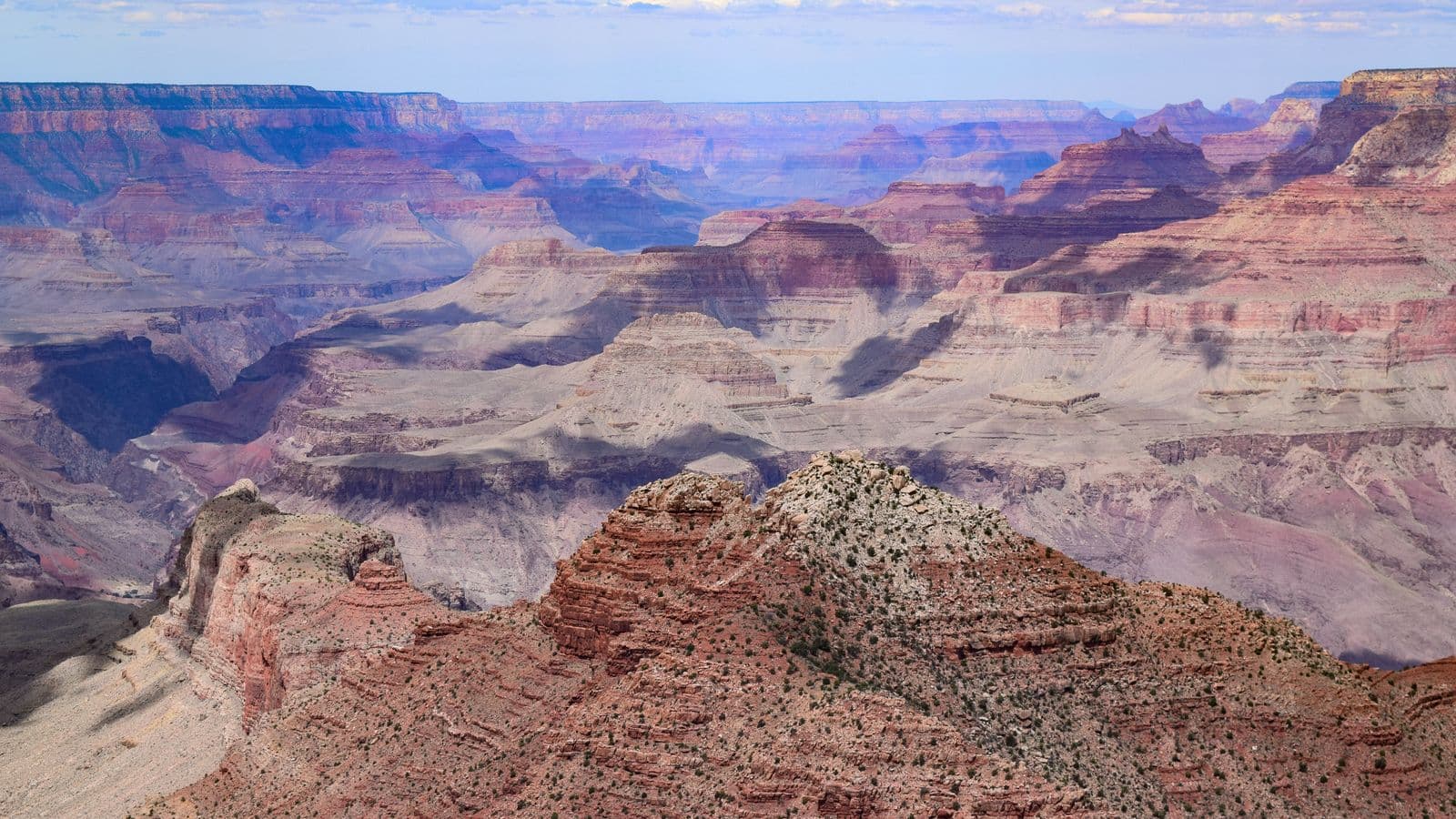 Layered red and purple buttes of the Grand Canyon stretch to the horizon beneath a hazy blue sky.