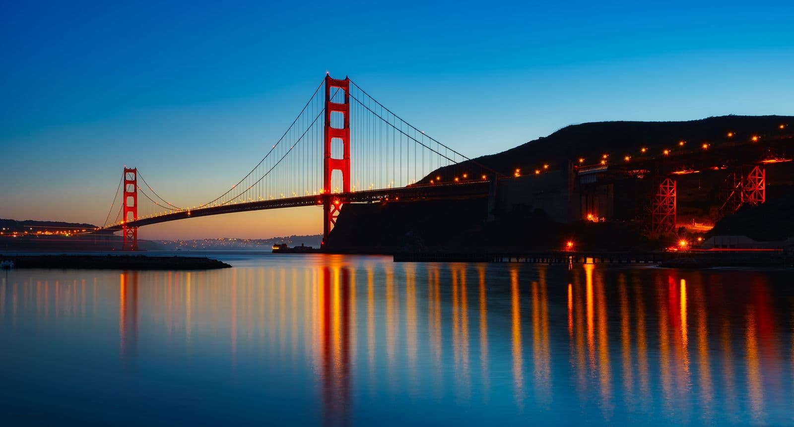 The Golden Gate Bridge glows at dusk, its lights reflecting in the calm water of San Francisco Bay.