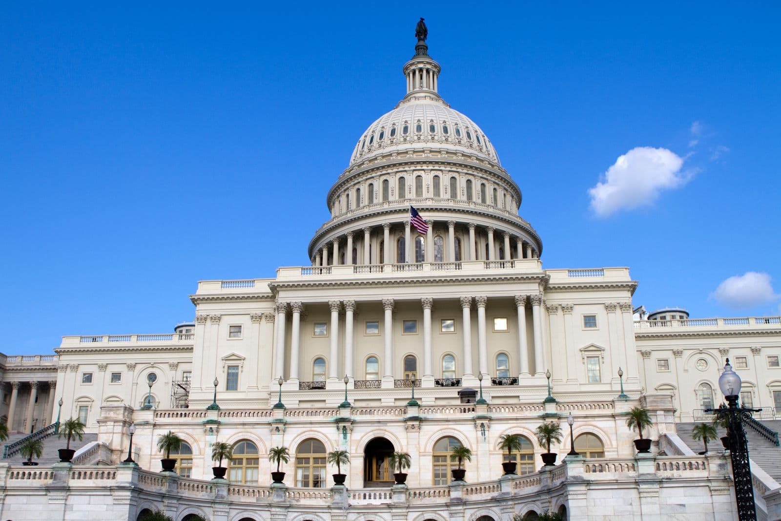 The west facade and dome of the United States Capitol rise under a bright blue sky with scattered clouds.