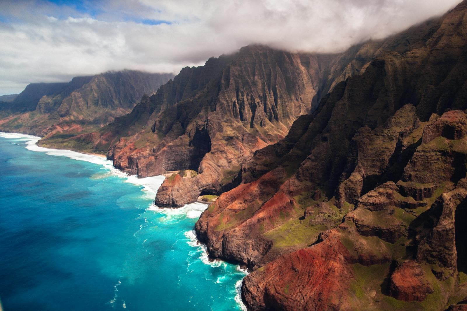 Emerald cliffs of Kauai's Na Pali Coast plunge into turquoise surf in a sweeping aerial view.