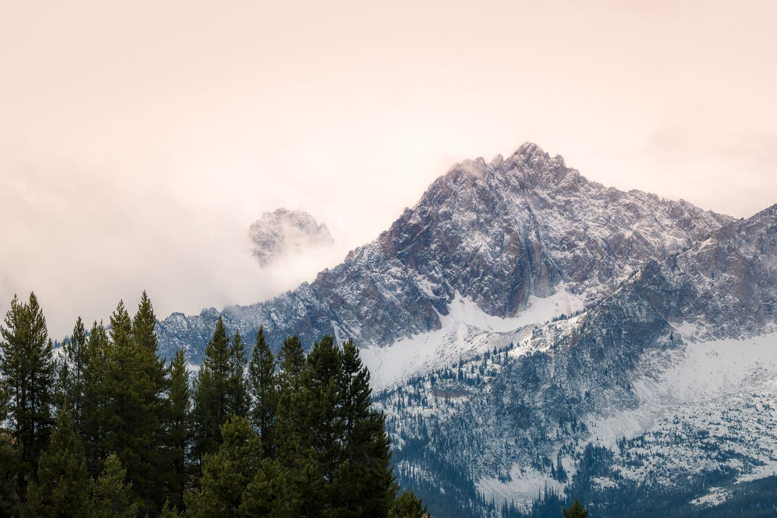 A snow-dusted Sawtooth peak near Stanley, Idaho rises through clouds into a soft pink sky above pines.