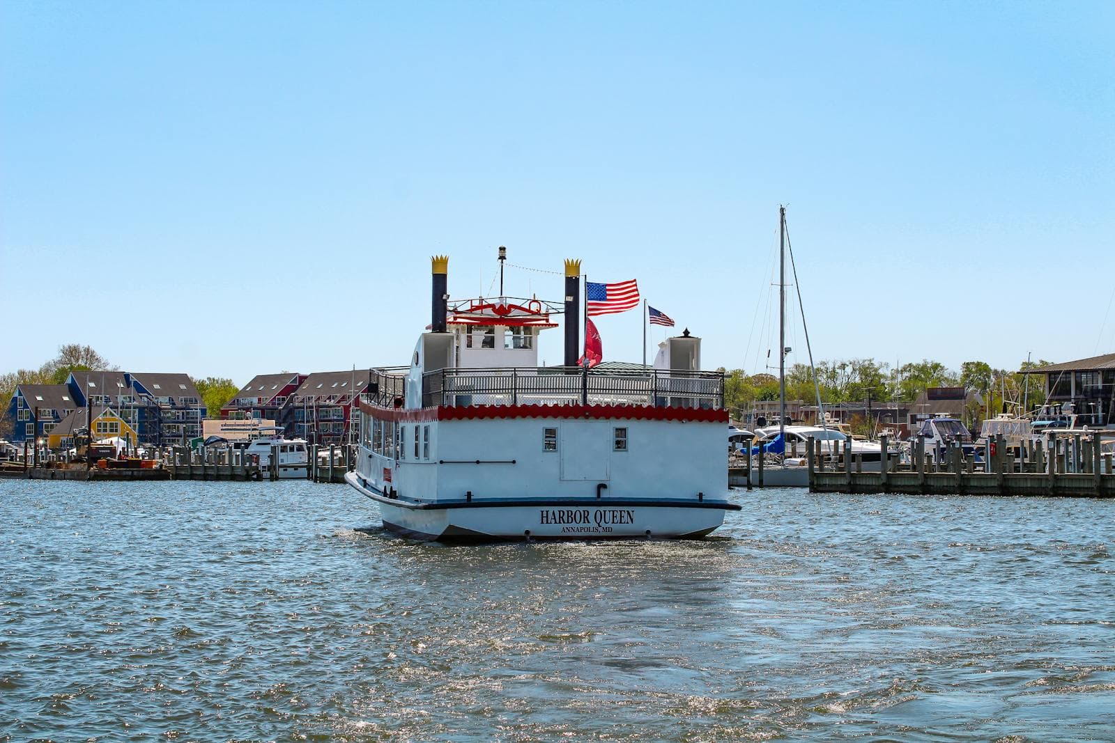 The Harbor Queen riverboat flying an American flag glides across Annapolis harbor with waterfront homes and sailboat masts along the shore.