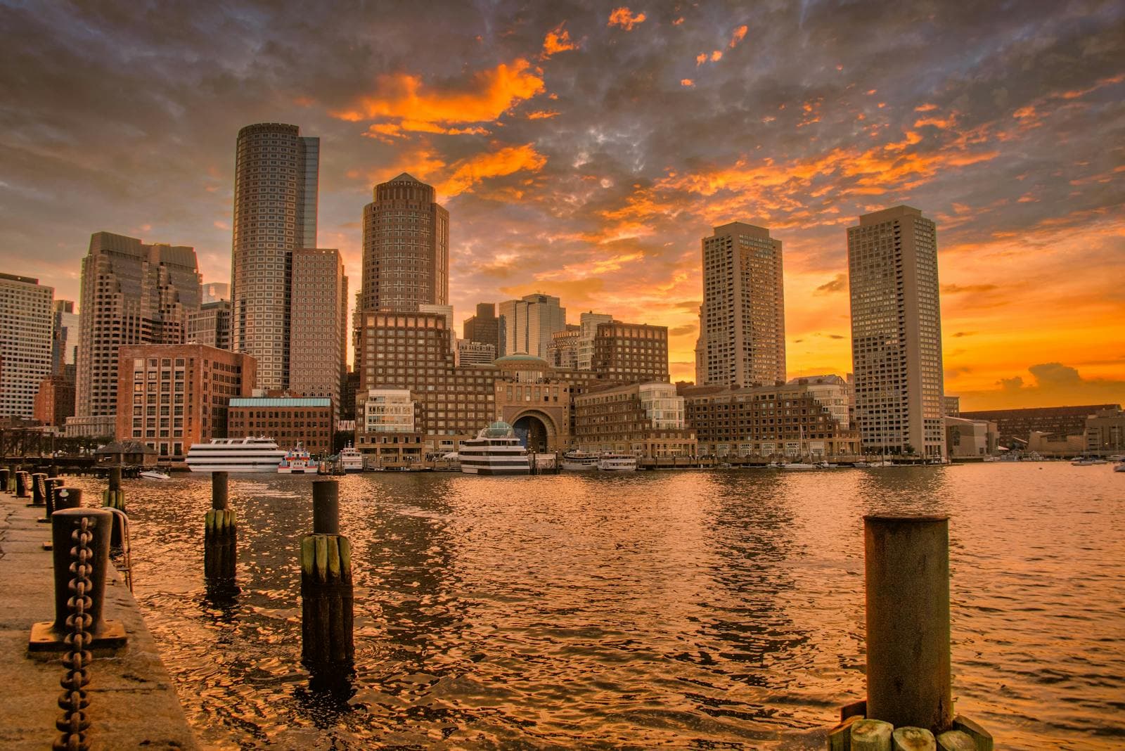 The Boston skyline glows at sunset with skyscrapers reflecting in the calm harbor waters.