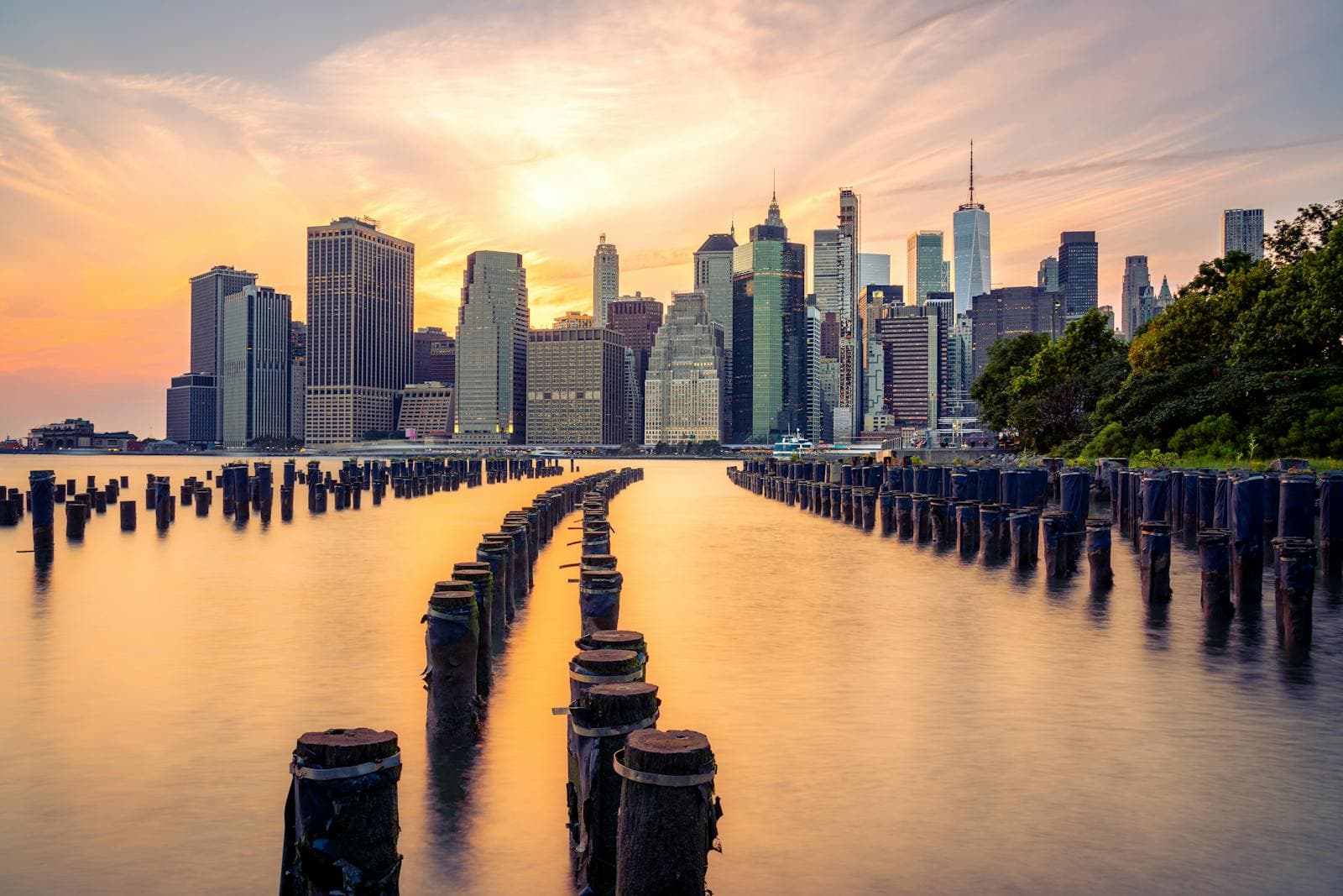 The Manhattan skyline glows under a warm sunset sky viewed across the East River from Brooklyn Bridge Park.
