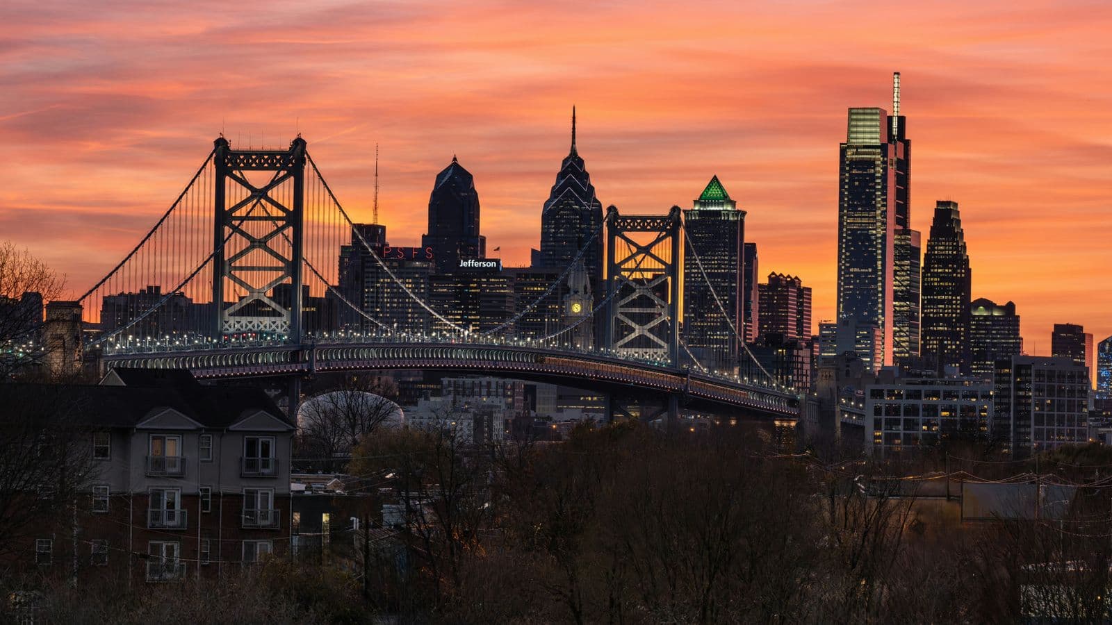 The Philadelphia skyline and Ben Franklin Bridge are silhouetted against a warm orange sunset over the Delaware River.