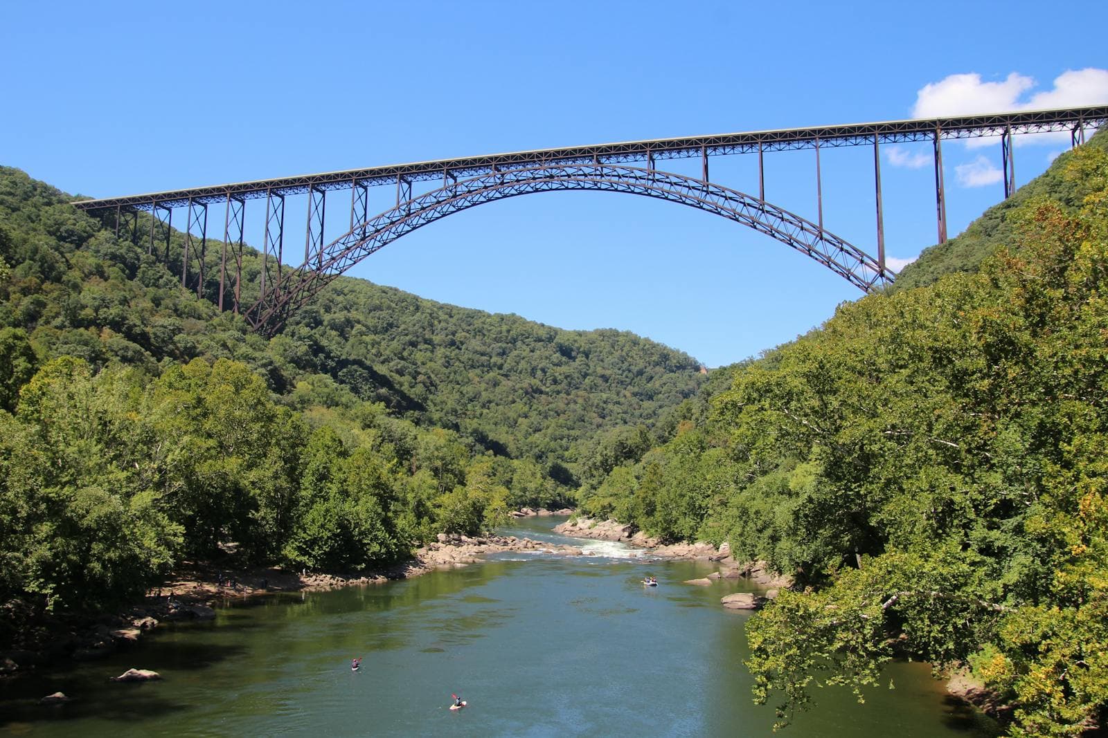 The steel arch of the New River Gorge Bridge spans high above a green forested gorge with rafters floating on the river below.